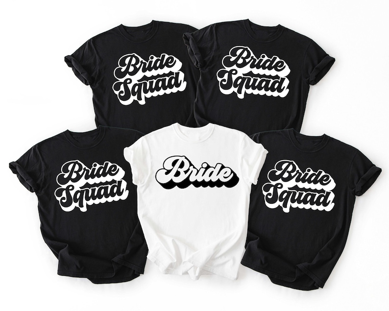 May include: Five t-shirts, four black and one white, with the words "Bride Squad" and "Bride" in a retro font. The black shirts have "Bride Squad" in white. The white shirt has "Bride" in black.