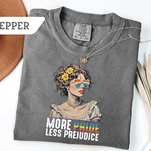May include: A gray t-shirt featuring a graphic of a woman wearing heart-shaped rainbow sunglasses and yellow flowers in her hair. The text "MORE PRIDE LESS PREJUDICE" is printed below the graphic. A silver necklace is draped across the shirt.