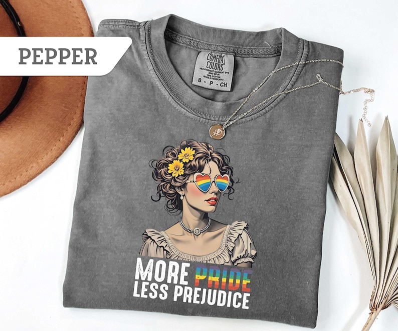 May include: A gray t-shirt featuring a graphic of a woman wearing heart-shaped rainbow sunglasses and yellow flowers in her hair. The text "MORE PRIDE LESS PREJUDICE" is printed below the graphic. A silver necklace is draped across the shirt.