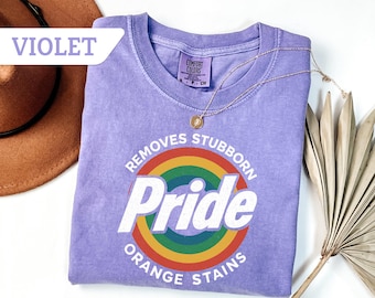 May include: A violet Comfort Colors t-shirt with a rainbow graphic that says "Removes Stubborn Pride Orange Stains." A gold necklace with a pendant is draped across the shirt. A brown hat is in the upper left corner.