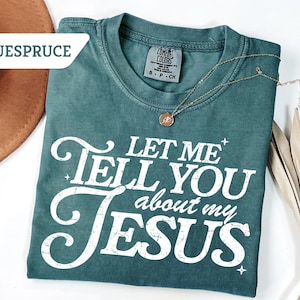 Puede incluir: Camiseta de color verde azulado con el texto blanco "LET ME TELL YOU about my JESUS". Un collar dorado con un colgante descansa sobre la camiseta. Un sombrero marr&oacute;n y hojas de palma secas en el fondo.