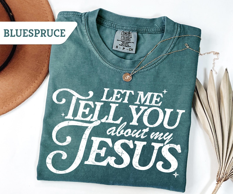 Puede incluir: Camiseta de color verde azulado con el texto blanco "LET ME TELL YOU about my JESUS". Un collar dorado con un colgante descansa sobre la camiseta. Un sombrero marr&oacute;n y hojas de palma secas en el fondo.