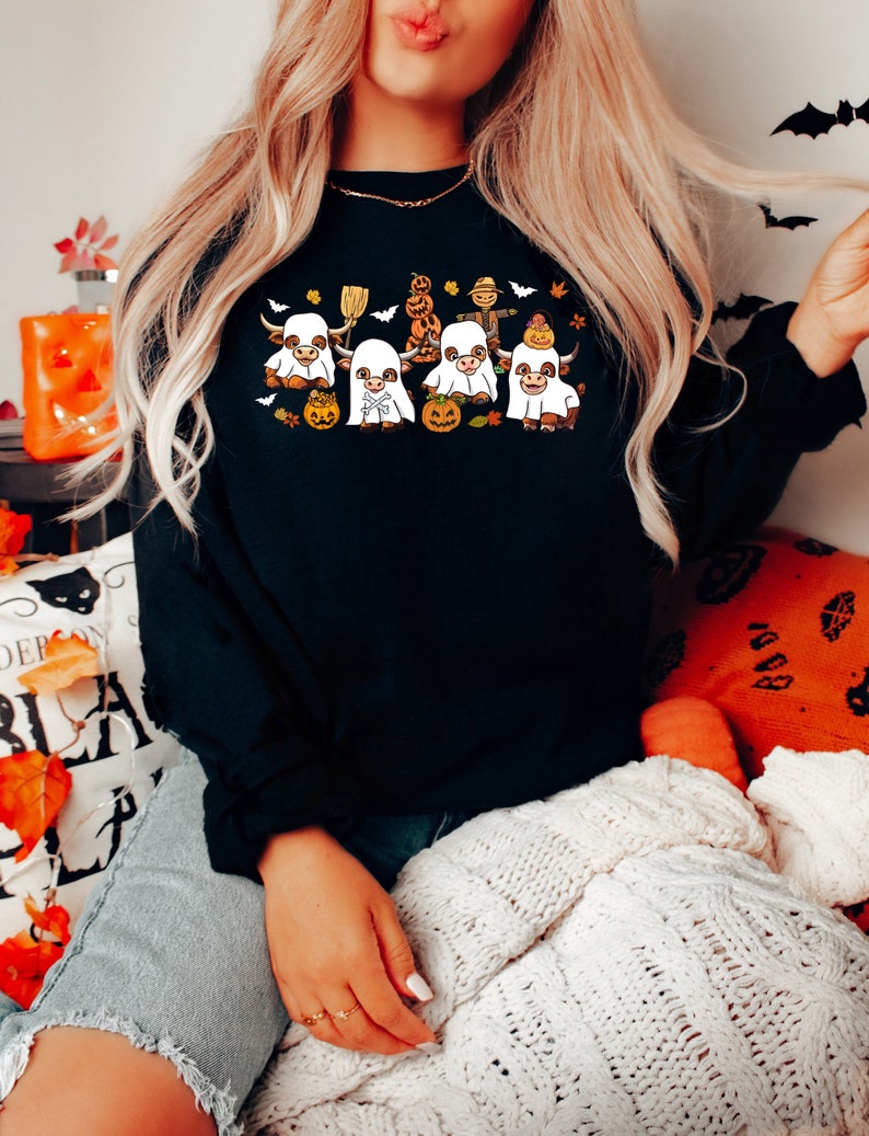 Op de afbeelding: Zwart sweatshirt met een afbeelding van vijf cartoonkoeien verkleed als spoken voor Halloween. De koeien dragen witte spokenkostuums met oranje pompoenen en vleermuizen eromheen.