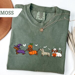 May include: Moss green t-shirt featuring a Halloween-themed dachshund design. The graphic includes four dachshunds dressed as a witch, pumpkin, mummy, and holding a pumpkin. The shirt has a crew neck and short sleeves.