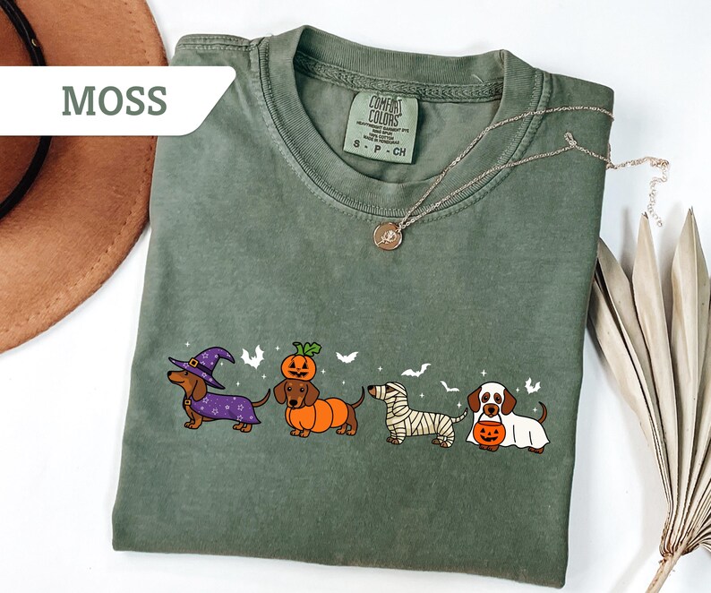 May include: Moss green t-shirt featuring a Halloween-themed dachshund design. The graphic includes four dachshunds dressed as a witch, pumpkin, mummy, and holding a pumpkin. The shirt has a crew neck and short sleeves.