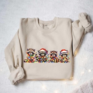 May include: A beige sweatshirt with a graphic of four raccoons wearing Santa hats and holding Christmas lights. The raccoons are surrounded by colorful Christmas lights.