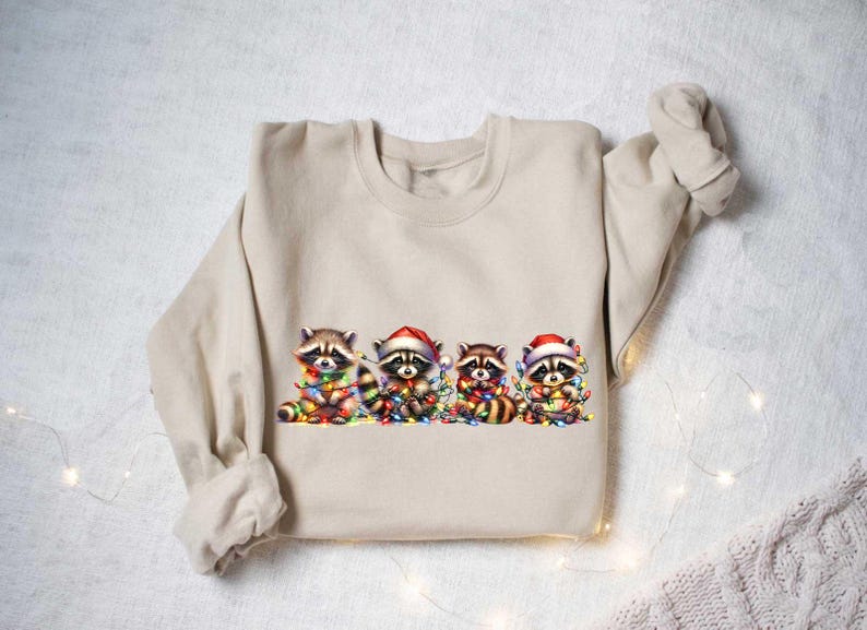 May include: A beige sweatshirt with a graphic of four raccoons wearing Santa hats and holding Christmas lights. The raccoons are surrounded by colorful Christmas lights.