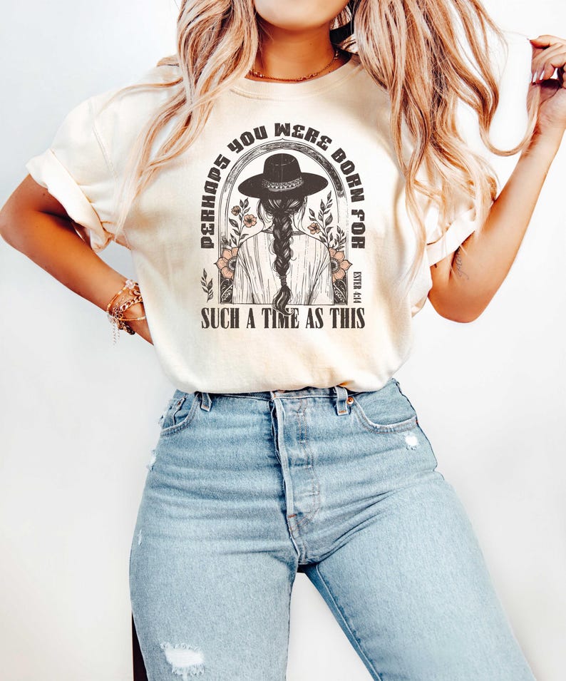 May include: A light beige t-shirt with a graphic print of a woman wearing a cowboy hat and the text "Perhaps you were born for such a time as this".