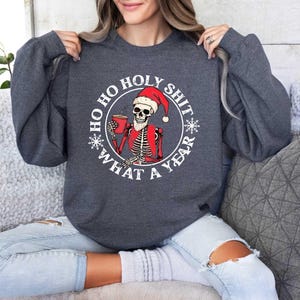 May include: A dark grey sweatshirt with a white circle graphic featuring a skeleton wearing a Santa hat and holding a cup of coffee. The text around the graphic reads "Ho Ho Holy Shit What A Year".