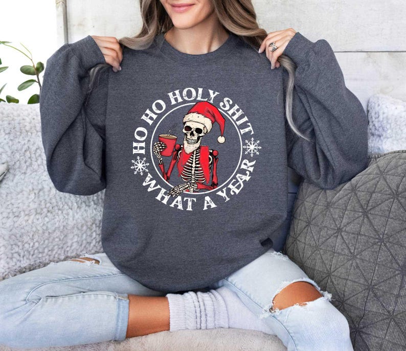 May include: A dark grey sweatshirt with a white circle graphic featuring a skeleton wearing a Santa hat and holding a cup of coffee. The text around the graphic reads "Ho Ho Holy Shit What A Year".