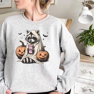 Puede incluir: Una sudadera gris con un gr&aacute;fico de un mapache vestido con un disfraz de fantasma y sosteniendo una bebida rosa. El mapache est&aacute; rodeado de dos calabazas naranjas con caras de jack-o'-lantern.