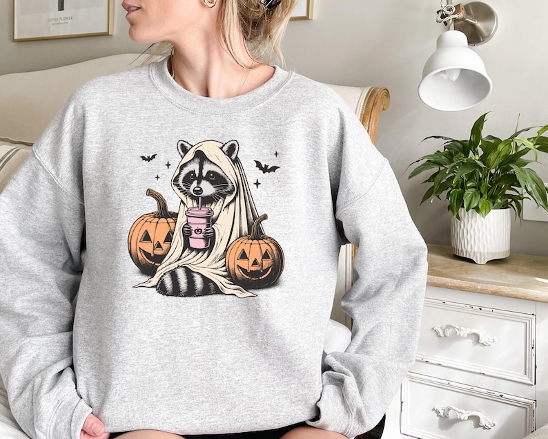 Puede incluir: Una sudadera gris con un gr&aacute;fico de un mapache vestido con un disfraz de fantasma y sosteniendo una bebida rosa. El mapache est&aacute; rodeado de dos calabazas naranjas con caras de jack-o'-lantern.