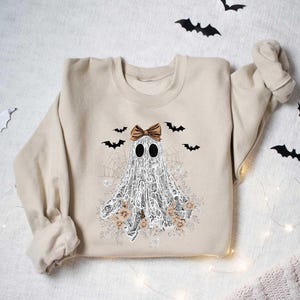Peut inclure: Un sweat-shirt beige avec un fant&ocirc;me blanc portant un n&oelig;ud brun et entour&eacute; de chauves-souris et de fleurs. Le fant&ocirc;me est en dentelle et a un motif de toile d'araign&eacute;e.