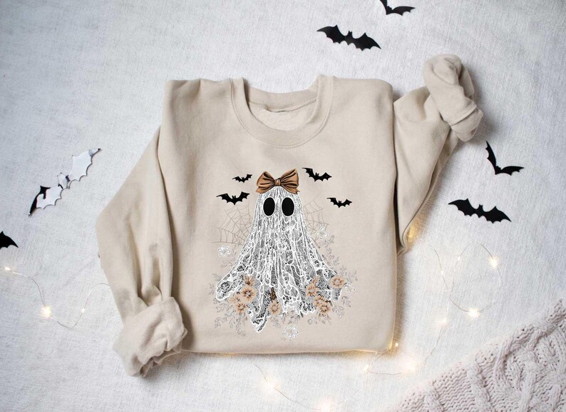Peut inclure: Un sweat-shirt beige avec un fant&ocirc;me blanc portant un n&oelig;ud brun et entour&eacute; de chauves-souris et de fleurs. Le fant&ocirc;me est en dentelle et a un motif de toile d'araign&eacute;e.