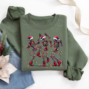 May include: Green Christmas sweater with a graphic of three skeletons wearing Santa hats and dancing with Christmas lights.