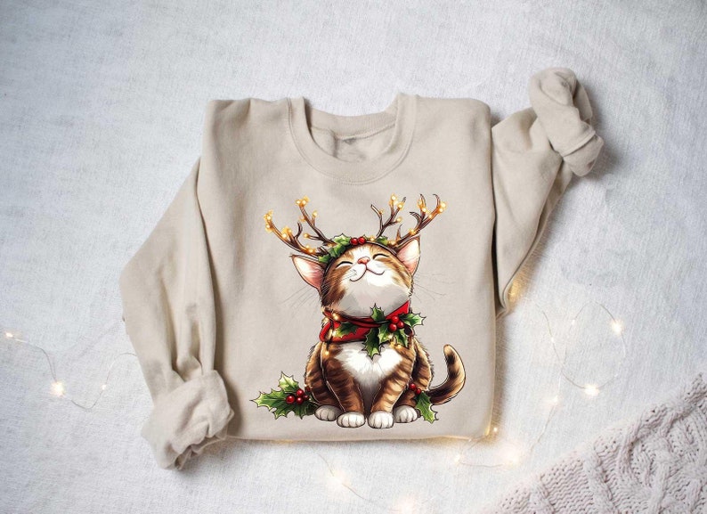 May include: A beige sweatshirt with a Christmas-themed graphic of a cat wearing a reindeer antler headband with lights and a red scarf with holly.