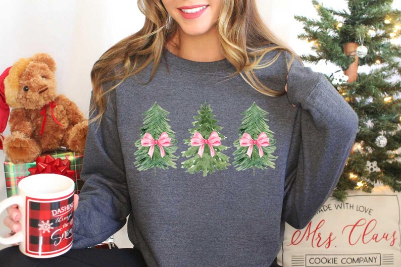 May include: A gray sweatshirt with a design of three watercolor Christmas trees with pink bows. The text "Mrs. Claus Cookie Company" is on the bottom of the sweatshirt.