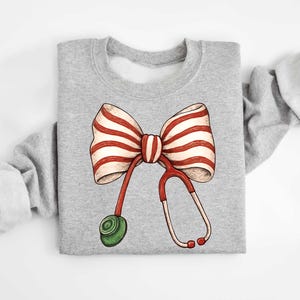 May include: A gray sweatshirt featuring a whimsical design of a red and white striped bow with a stethoscope. The stethoscope is red and white with a green earpiece. The design is centered on the front of the sweatshirt.