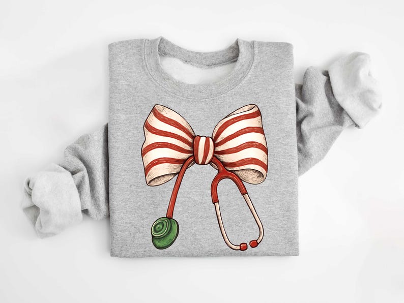 May include: A gray sweatshirt featuring a whimsical design of a red and white striped bow with a stethoscope. The stethoscope is red and white with a green earpiece. The design is centered on the front of the sweatshirt.