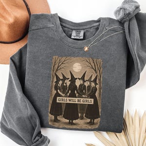 K&ouml;nnte beinhalten: Ein anthrazitfarbenes Sweatshirt mit einer Grafik von sechs Hexen in schwarzen Kleidern und spitzen H&uuml;ten, die sich an den H&auml;nden halten. Das Bild enth&auml;lt den Text "GIRLS WILL BE GIRLS" auf einem Banner. Ein brauner Hut und eine Halskette sind ebenfalls sichtbar.