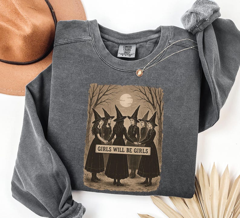 K&ouml;nnte beinhalten: Ein anthrazitfarbenes Sweatshirt mit einer Grafik von sechs Hexen in schwarzen Kleidern und spitzen H&uuml;ten, die sich an den H&auml;nden halten. Das Bild enth&auml;lt den Text "GIRLS WILL BE GIRLS" auf einem Banner. Ein brauner Hut und eine Halskette sind ebenfalls sichtbar.