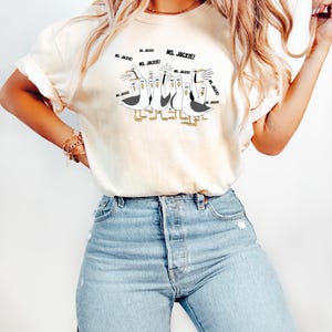 May include: Cream-colored t-shirt featuring a cartoon illustration of seagulls with the text "MS. JACKIE!" in speech bubbles. The seagulls are depicted in black, white, and yellow, with the text in black. The shirt is paired with light blue denim jeans.