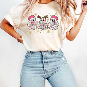 Puede incluir: Una camiseta blanca con un dise&ntilde;o festivo de tres fantasmas con gorros de Pap&aacute; Noel y sosteniendo regalos. Los fantasmas est&aacute;n decorados con patrones florales rosas y verdes. El dise&ntilde;o incluye copos de nieve y purpurina dorada.