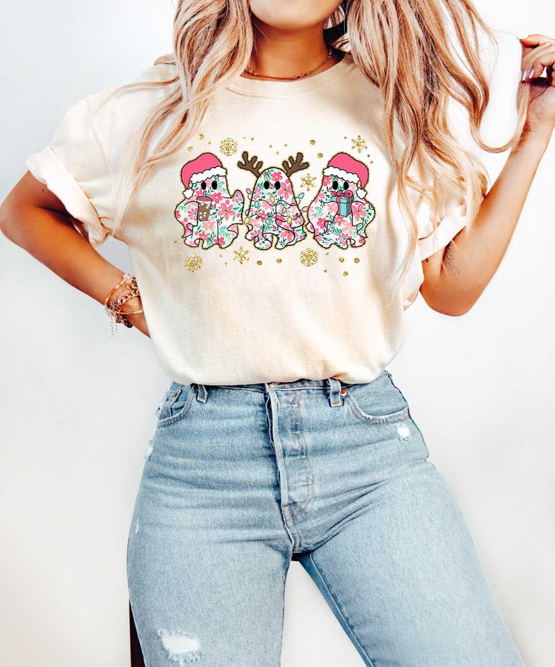 Puede incluir: Una camiseta blanca con un dise&ntilde;o festivo de tres fantasmas con gorros de Pap&aacute; Noel y sosteniendo regalos. Los fantasmas est&aacute;n decorados con patrones florales rosas y verdes. El dise&ntilde;o incluye copos de nieve y purpurina dorada.