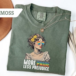 May include: Moss green t-shirt featuring a graphic of a woman with rainbow heart-shaped sunglasses and the text "MORE PRIDE LESS PREJUDICE". The shirt has a crew neck and short sleeves. A gold necklace is draped across the shirt.