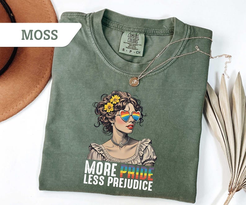 May include: Moss green t-shirt featuring a graphic of a woman with rainbow heart-shaped sunglasses and the text "MORE PRIDE LESS PREJUDICE". The shirt has a crew neck and short sleeves. A gold necklace is draped across the shirt.