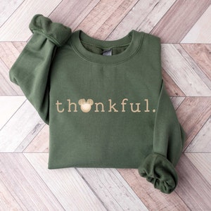 May include: A green sweatshirt with the word "thankful" in gold lettering and a gold Mickey Mouse head graphic.