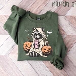 Puede incluir: Una sudadera verde con un estampado gr&aacute;fico de un mapache vestido de fantasma y sosteniendo una bebida rosa. El mapache est&aacute; rodeado de calabazas y murci&eacute;lagos. El texto "MILITARY GREEN" est&aacute; impreso en blanco en la parte superior de la imagen.