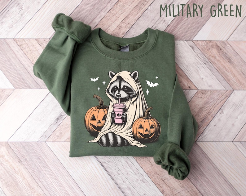 Puede incluir: Una sudadera verde con un estampado gr&aacute;fico de un mapache vestido de fantasma y sosteniendo una bebida rosa. El mapache est&aacute; rodeado de calabazas y murci&eacute;lagos. El texto "MILITARY GREEN" est&aacute; impreso en blanco en la parte superior de la imagen.