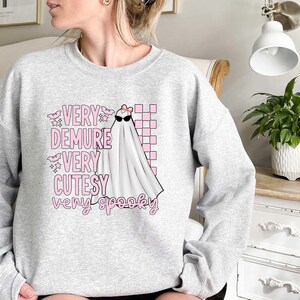 May include: A gray sweatshirt with a pink and white graphic of a ghost wearing sunglasses and a bow. The text on the sweatshirt reads "Very Demure Very Cutesy Very Spooky."