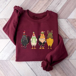 May include: A maroon sweatshirt with a festive design of four chickens wearing Christmas hats and lights. The chickens are wearing different colored hats and have different expressions.