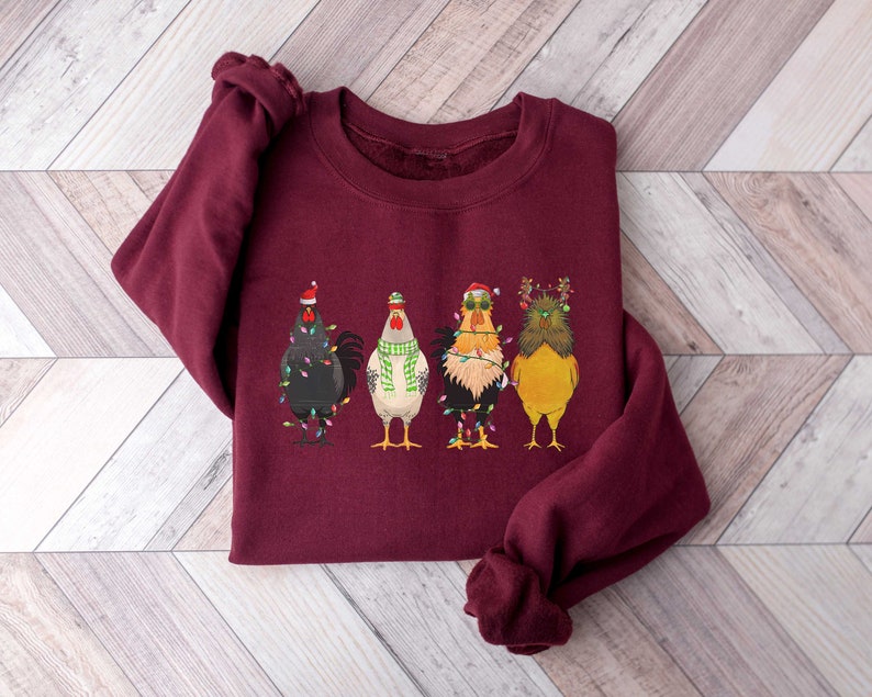 May include: A maroon sweatshirt with a festive design of four chickens wearing Christmas hats and lights. The chickens are wearing different colored hats and have different expressions.