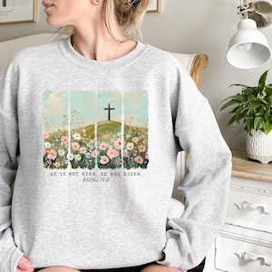 May include: A gray sweatshirt with a floral design and a cross on a hill. The text "He is not here. He has risen. Matthew 28:6" is printed below the design.