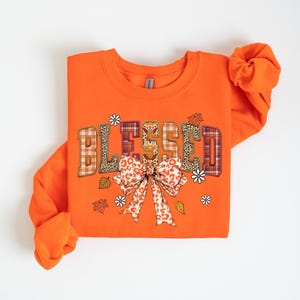 May include: Orange sweatshirt with the word "BLESSED" in a decorative font. The letters have plaid, leopard print, and floral patterns. A large bow with a similar pattern is below the word, with leaves and flowers. A fall-themed apparel item.