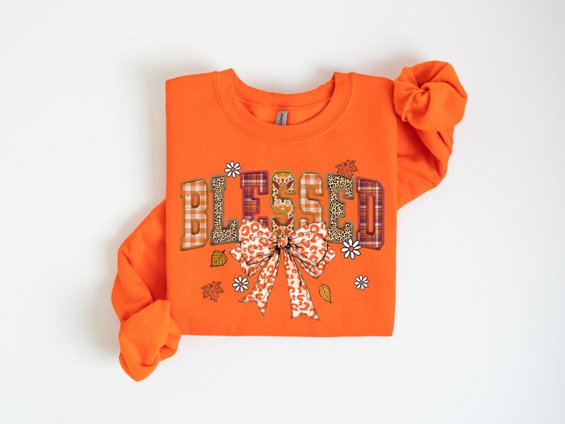 May include: Orange sweatshirt with the word "BLESSED" in a decorative font. The letters have plaid, leopard print, and floral patterns. A large bow with a similar pattern is below the word, with leaves and flowers. A fall-themed apparel item.