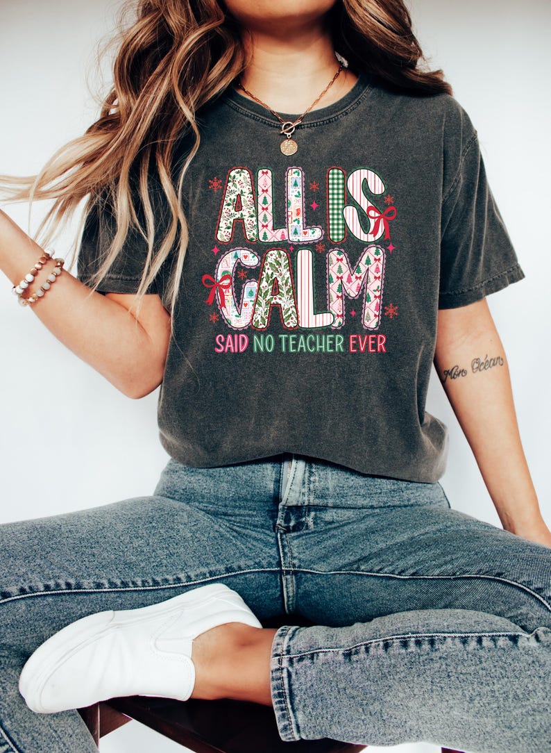 May include: Dark gray t-shirt with the text "ALL IS CALM SAID NO TEACHER EVER" in a colorful, festive design. The design incorporates Christmas tree and bow graphics. A casual, comfortable tee for teachers.