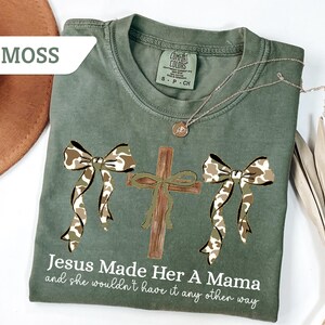 May include: Moss green t-shirt with a cross and two bows in a camouflage pattern. The text "Jesus Made Her A Mama and she wouldn't have it any other way" is printed on the shirt. A gold necklace is draped across the shirt.