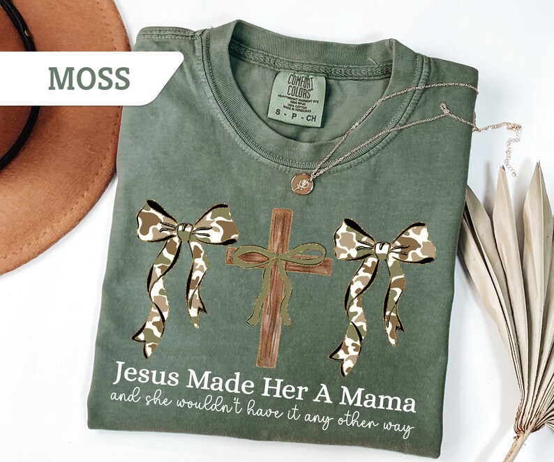 May include: Moss green t-shirt with a cross and two bows in a camouflage pattern. The text "Jesus Made Her A Mama and she wouldn't have it any other way" is printed on the shirt. A gold necklace is draped across the shirt.