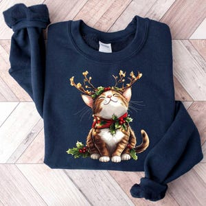 May include: A navy blue sweatshirt with a graphic of a brown and white cat wearing a red and green reindeer antler headband. The cat is smiling and has a red and green bow around its neck.