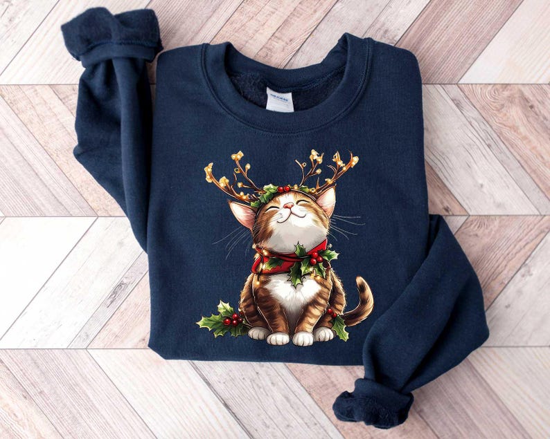 May include: A navy blue sweatshirt with a graphic of a brown and white cat wearing a red and green reindeer antler headband. The cat is smiling and has a red and green bow around its neck.