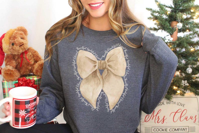 May include: A gray sweatshirt with a large beige bow design. The bow is surrounded by the words "Thankful, Grateful, Blessed" repeated in a circle.