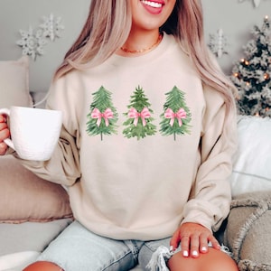 May include: A beige sweatshirt with a watercolor design of three Christmas trees with pink bows.