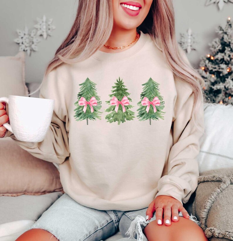 May include: A beige sweatshirt with a watercolor design of three Christmas trees with pink bows.