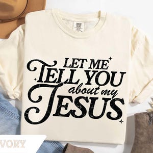 Puede incluir: Camiseta de color marfil con el texto "LET ME TELL YOU about my JESUS" en negro, con una fuente estilizada. La camiseta es de la marca Comfort Colors. La camiseta se muestra con un sombrero, vaqueros y botas.