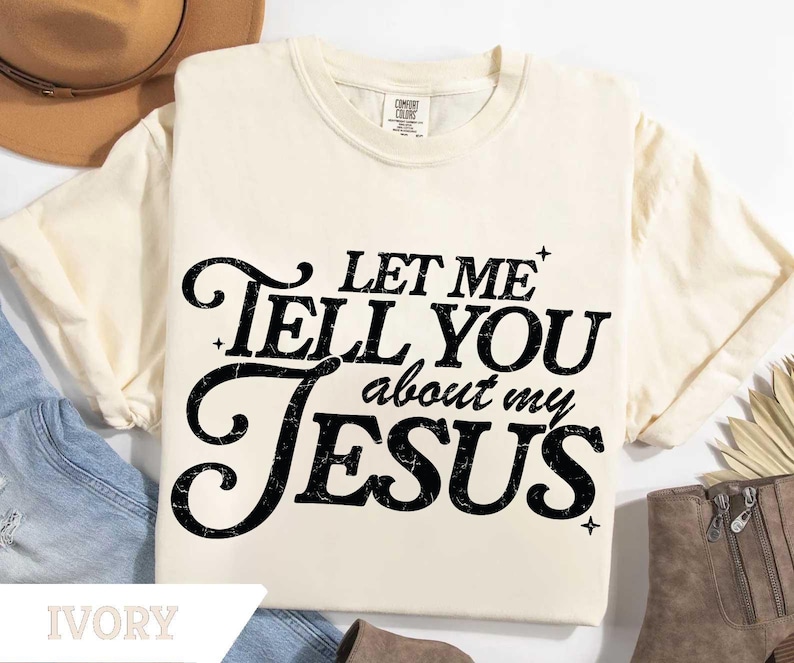Puede incluir: Camiseta de color marfil con el texto "LET ME TELL YOU about my JESUS" en negro, con una fuente estilizada. La camiseta es de la marca Comfort Colors. La camiseta se muestra con un sombrero, vaqueros y botas.
