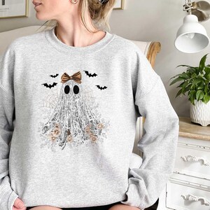 Peut inclure: Un sweat-shirt gris avec un fant&ocirc;me blanc portant un n&oelig;ud brun et entour&eacute; de chauves-souris et de fleurs. Le fant&ocirc;me est fait de dentelle.
